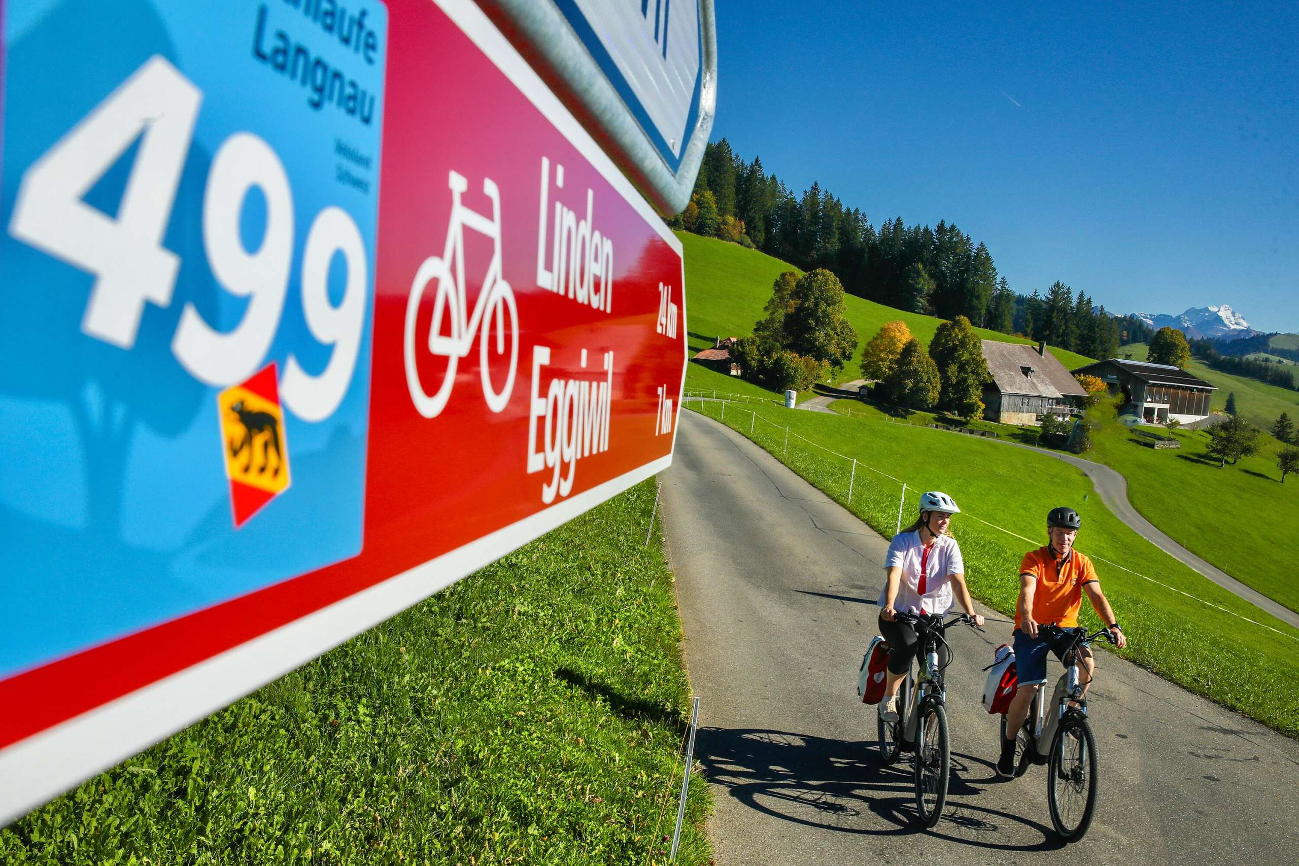 Zwei E-Bike Fahrer unterwegs auf der Herzschlaufe Langnau Nr. 499. Im Vordergrund der Wegweiser. Im Hintergrund Berge und Bauernhof.